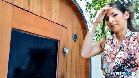Sexy brunette babe shades eyes smiling seductively outside wooden sauna in revealing floral top