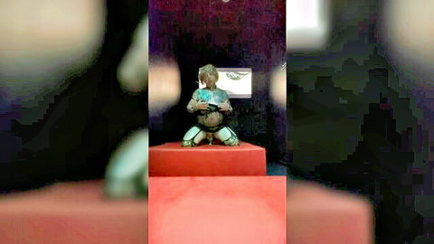 Pierced sissy in chains, seated in green fetish outfit on pedestal