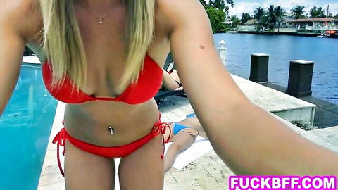 Blonde babe in red string bikini shows off huge tits and navel piercing poolside with blue bikini friend