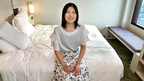 Beautiful Japanese woman in white blouse and floral skirt sitting on bed, smiling seductively