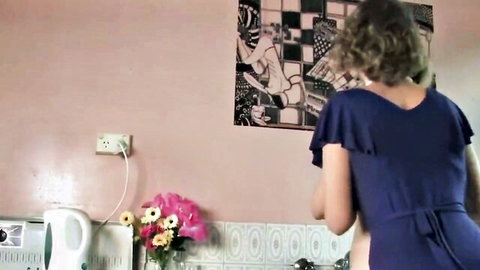 Curly-haired woman in blue dress turns away in kitchen, teasing lesbian mom seduction