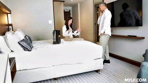 Asian teen in white blouse sits smiling on hotel bed with black stud in suit