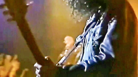 Curly haired rocker in shiny blue jacket shredding guitar on stage with mic stand