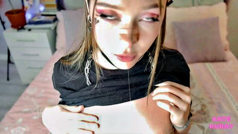 Seductive e-girl with heavy makeup, choker and chains teases in black top on pink bed