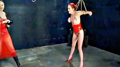 Redhead bound in chains with exposed tits and shaved pussy dominated by latex mistress in BDSM dungeon