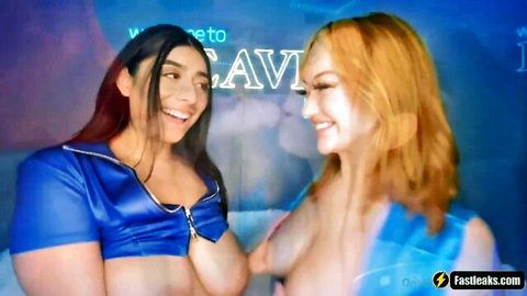 Two busty women in shiny blue tops smiling close-up, teasing cleavage