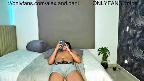 Muscular blond guy in gray briefs on bed, holding phone - Alex and Dani gay tease