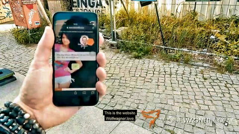 Hand holding iPhone displaying young brunette woman on cam chat screen with Wolfe Wagner watermark outdoors