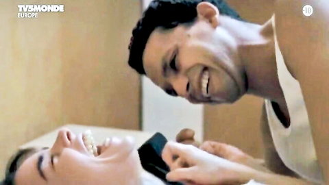 Playful couple laughing intimately in bed, man leaning over smiling woman holding phone