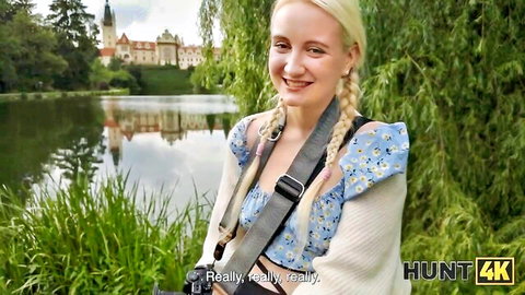Petite blonde with pigtails smiles teasingly outdoors in park by lake with castle