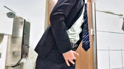 Suited East Asian man teasing at public restroom urinal with hand near zipper bulge
