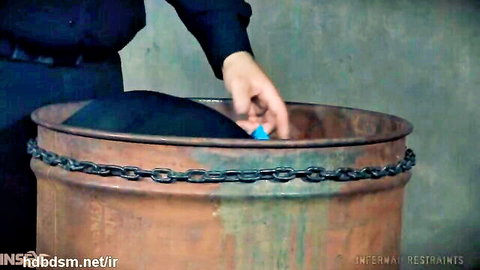Gloved hand retrieves small blue toy from chained rusty barrel in BDSM slave dungeon