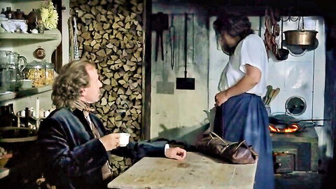 Clothed man sips tea at rustic kitchen table while woman in white blouse and blue skirt stands nearby
