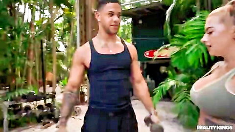 Busty blonde in gray sports bra flirts with muscular tattooed black stud in outdoor gym
