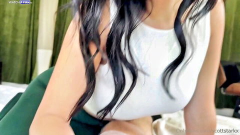 Cami Strella in white tank top showing deep cleavage, long black hair, on bed