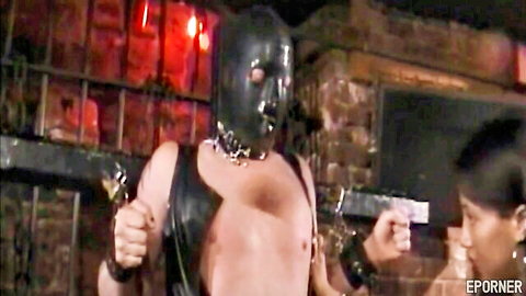 Muscular BDSM slave in shiny latex mask and chain harness poses wildly for Asian femdom mistress in dungeon