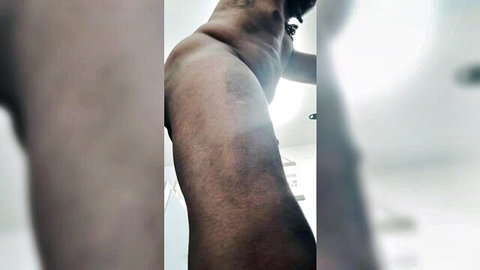 Tattooed dark-skinned man stands nude in bathroom showing side profile of his erect cock and muscular body