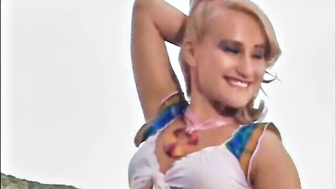 Smiling blonde babe posing with arms raised in low-cut white top and colorful straps