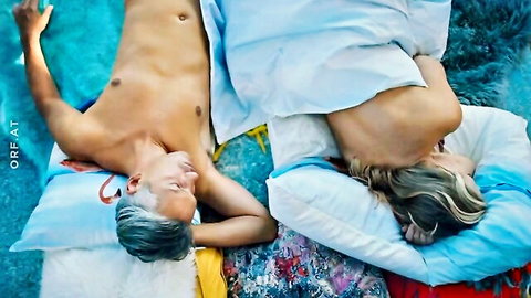 Topless gray-haired man and nude blonde woman lying naked on colorful bedroom bed sheets