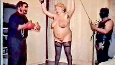 Nude blonde BBW with huge tits in BDSM scene, arms bound, leashed by masked dom, man with weights