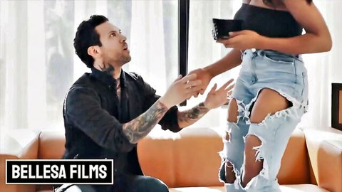 Tattooed man kneels proposing to curvy dark-skinned woman in ripped jeans and black top