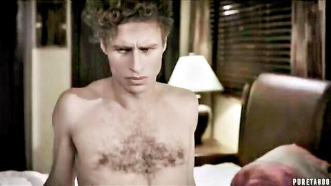 Shirtless young curly-haired man with hairy chest sits seriously on bedroom bed