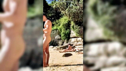 Young nude man standing on sunny beach with another nudist lounging nearby