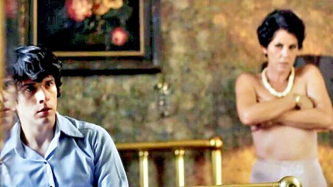 Young man stares at topless mature woman with bare tits and pearl necklace arms crossed in bedroom