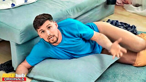 Horny guy with erect cock exposed lying on floor in blue t-shirt and orange shorts surprised expression