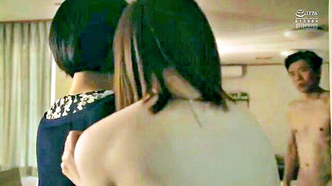 Shirtless Japanese man approaches two petite women from behind in dimly lit bedroom.