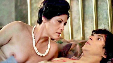 Topless busty MILF with pearl necklace leans over young stud's scratched bloody chest in rough sex aftermath