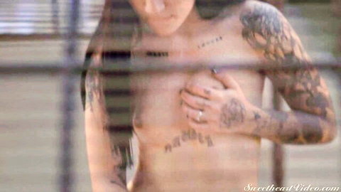 Goth Charlotte topless with tattoos, hand on breast behind bars