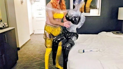 Redhead in yellow PVC bikini embraces plastic-wrapped sissy in black latex on hotel bed
