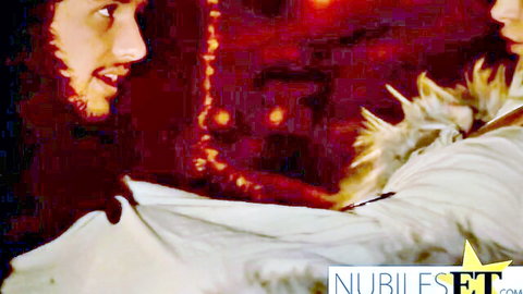 Bearded man stares intimately at blonde woman in feathered white top on red velvet background