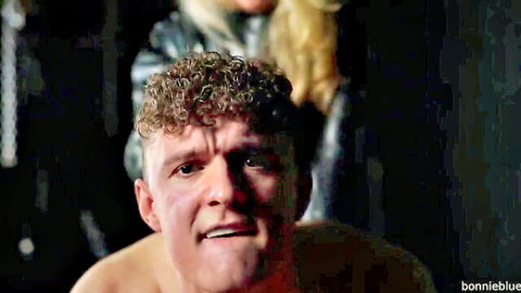 Blonde leather-clad domme towers over curly-haired shirtless guy gasping mouth open in strapon pegging domination