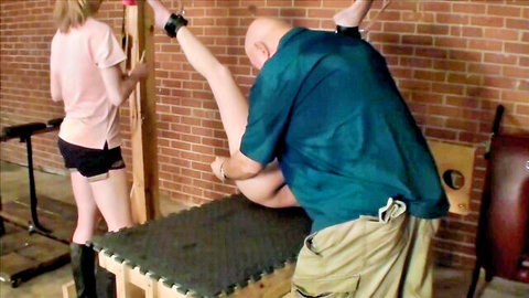 Blonde woman in tight shorts has leg cuffed high on bondage frame while bald man grips her thigh