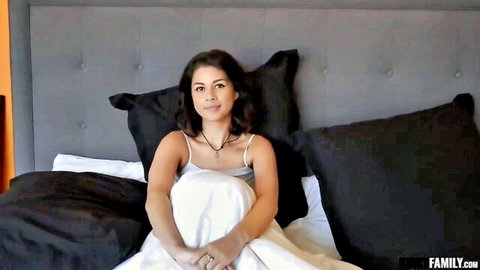 Cute brunette Latina girl in white tank top smiling on bed, holding sheet seductively