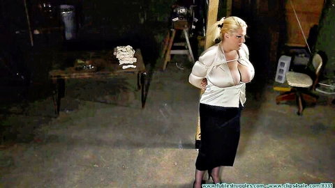 Busty blonde dominatrix bound with hands behind back in blouse and skirt