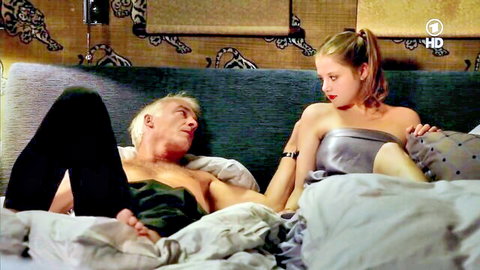 Jella Haase in silver top with older man on bed, teasing couple scene