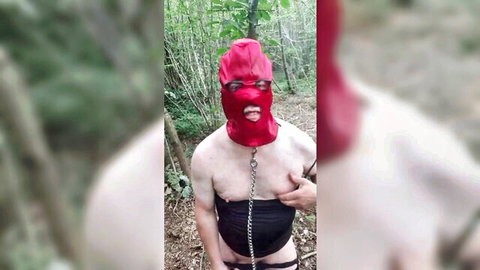 Red latex hooded sissy with chain collar and black crotch harness topless in forest
