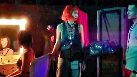 Petite redhead with small tits exposed topless in neon-lit dressing room with other nude petite girls