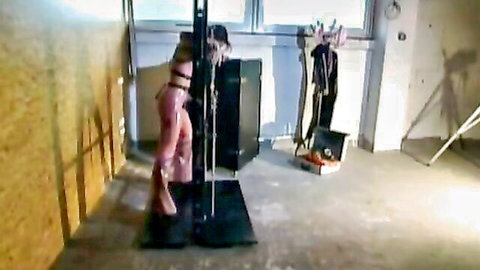 Pink latex clad woman bound spread-eagle on metal BDSM cross in dungeon teasing her ass