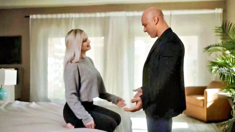 Blonde babe in gray crop top and leggings sits on bed facing bald suited man in bedroom