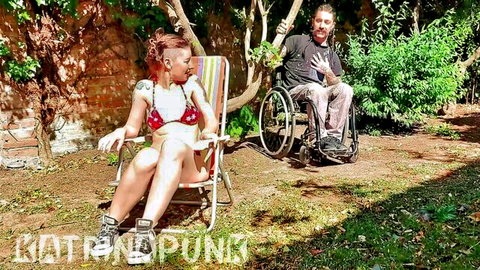 Redhead in pink bikini lounging seductively on chair next to smiling guy in wheelchair outdoors