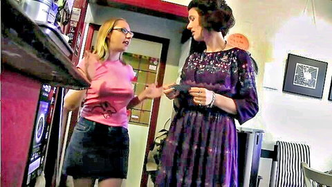 Blonde in pink top and denim skirt hands item to brunette in purple dress in teasing lesbian kitchen scene