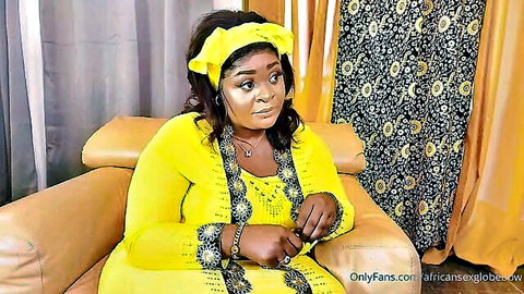Curvy ebony BBW MILF sitting on couch in yellow embroidered dress smiling seductively