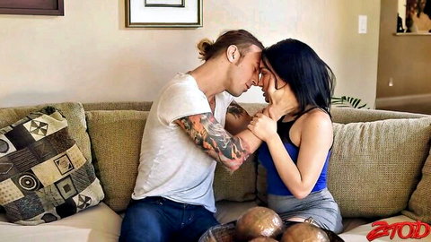 Tattooed guy with man bun passionately kissing skinny brunette in blue sports bra and gray skirt on couch