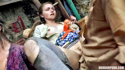 Distressed blonde woman in horror porn, exposed small breasts, holding doll, pants down