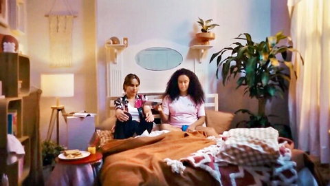 Two clothed women sitting on bed eating snacks in cozy bedroom