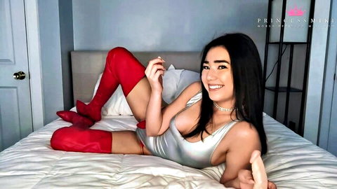 Seductive Asian woman in red thigh-high boots and silver bodysuit smiling on bed